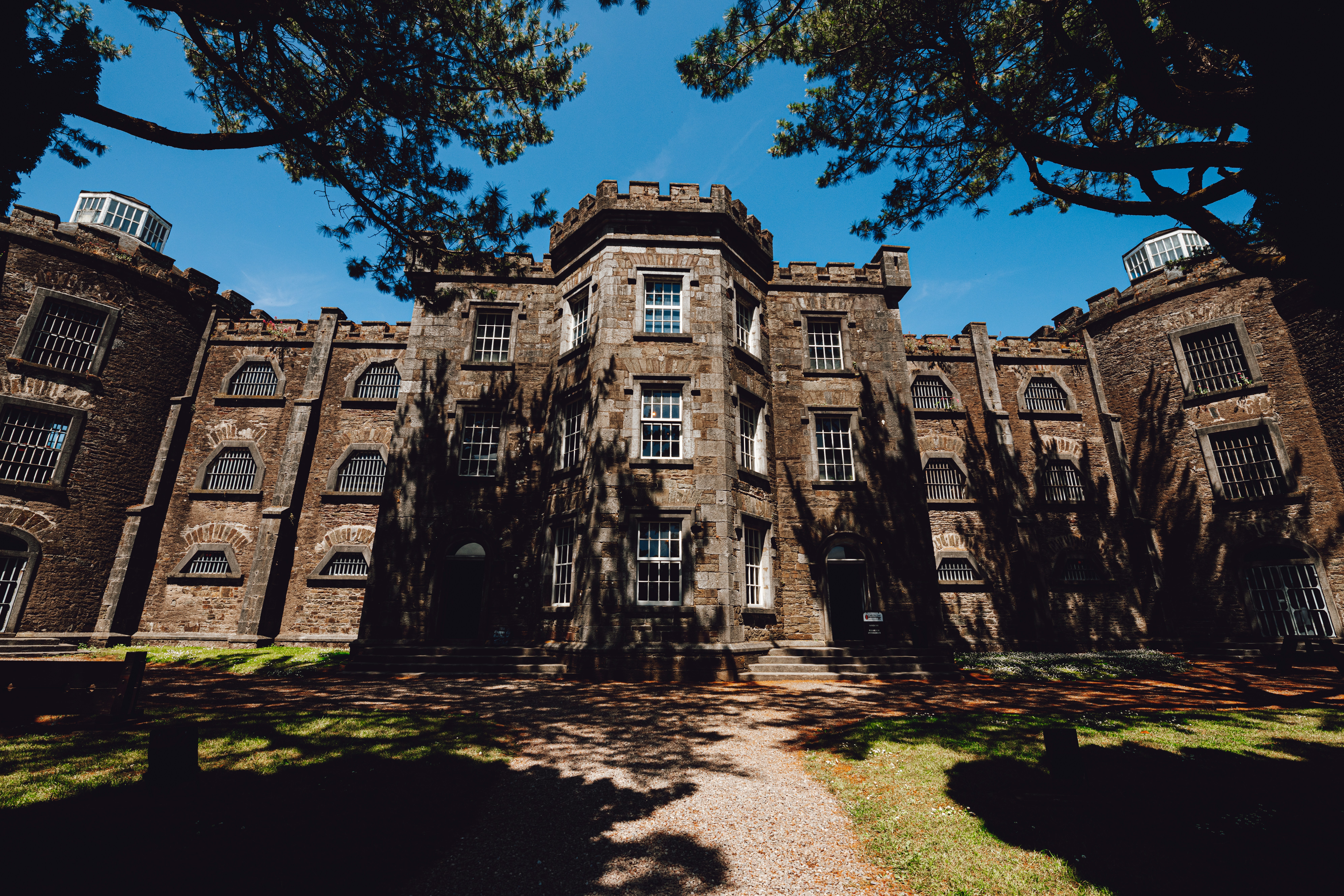 The castellated stone exterior of Cork City Gaol, with barred windows and tree shadows across the historic fa&ccedil;ade.
